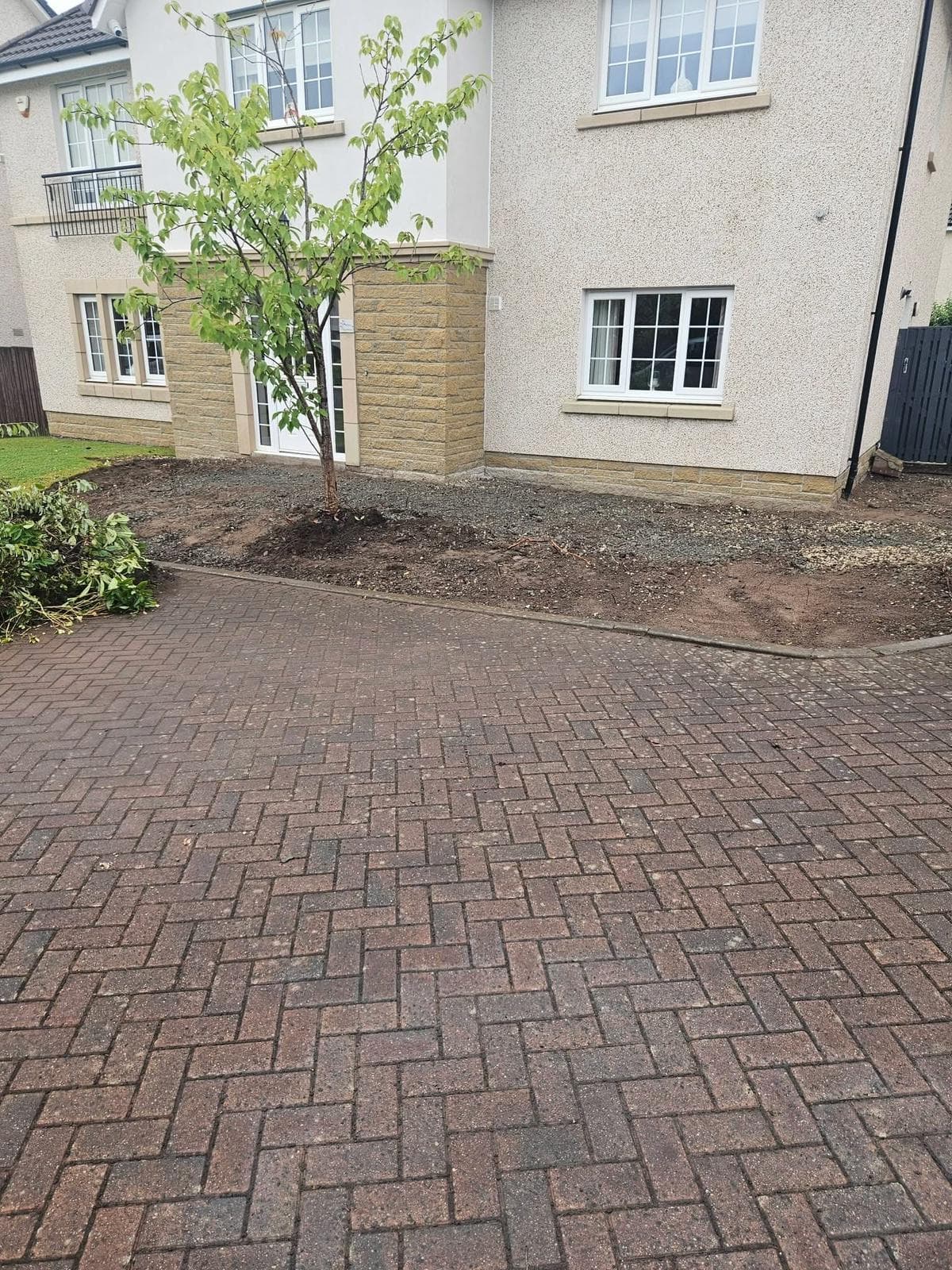 Wider view of the Dargavel frontage before work, showing builder stone, bare soil and prunings piled at the lawn edge